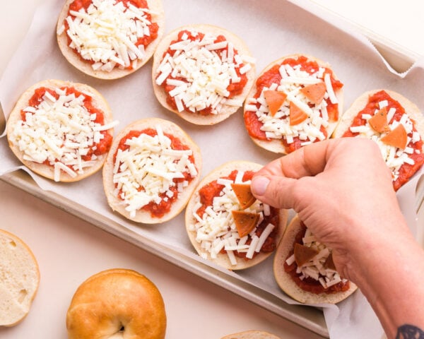 A hand puts vegan pepperoni pizza on mini bagels topped with pizza sauce and vegan mozzarella shreds. There are mini bagels sitting by the pan.