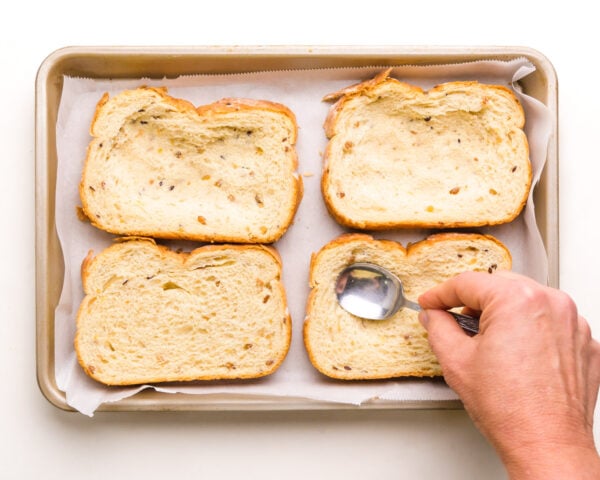 Four slices of bread are in a baking pan. A hand holds a spoon, pressing in the center to create a well in each slice of bread.
