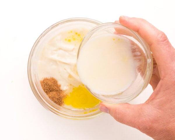 A hand holds a bowl with a "flour egg", pouring it into another bowl with yogurt and spices.