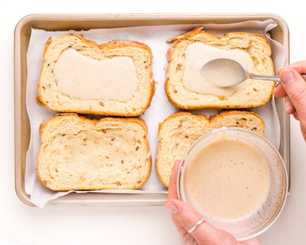 A hand holds a bowl of custard, and is using a spoon to distribute into four slices of bread in a baking pan.