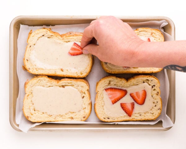 A hand is adding sliced strawberries on top of pieces of bread topped with yogurt custard in a baking pan.