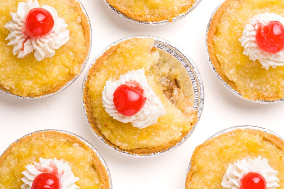 Looking down on pineapple tarts with whipped cream and cherries on top. One of them has a bite taken out.