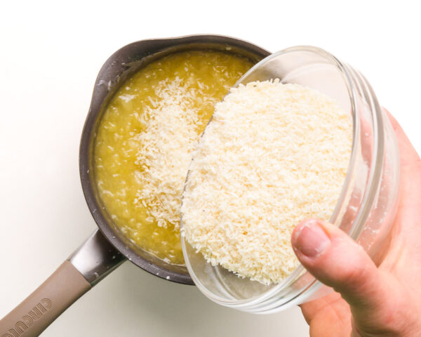 A hand holds a bowl of coconut flakes, pouring them into a saucepan with a pineapple mixture.