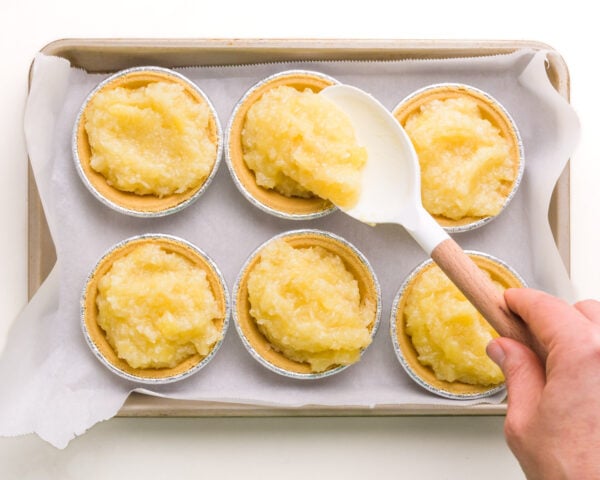 A hand holds a spatula, spooning pineapple filling into tarts in a baking pan.