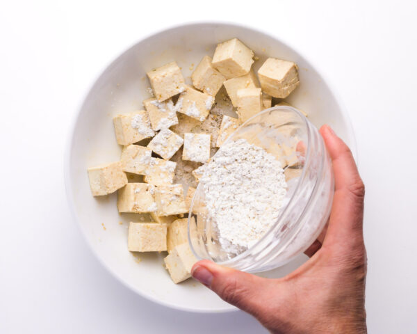 cornstarch is being spriled over tofu cubes in a bowl.