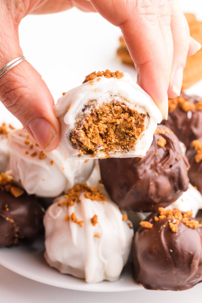 A hand holds a Biscoffe ball with a bite taken out. It hovers over a plate with more of the treats.