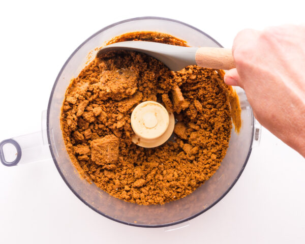 A hand holds a spatula, scraping down the sides of a food processor bowl with crumbled biscoff cookies in it.