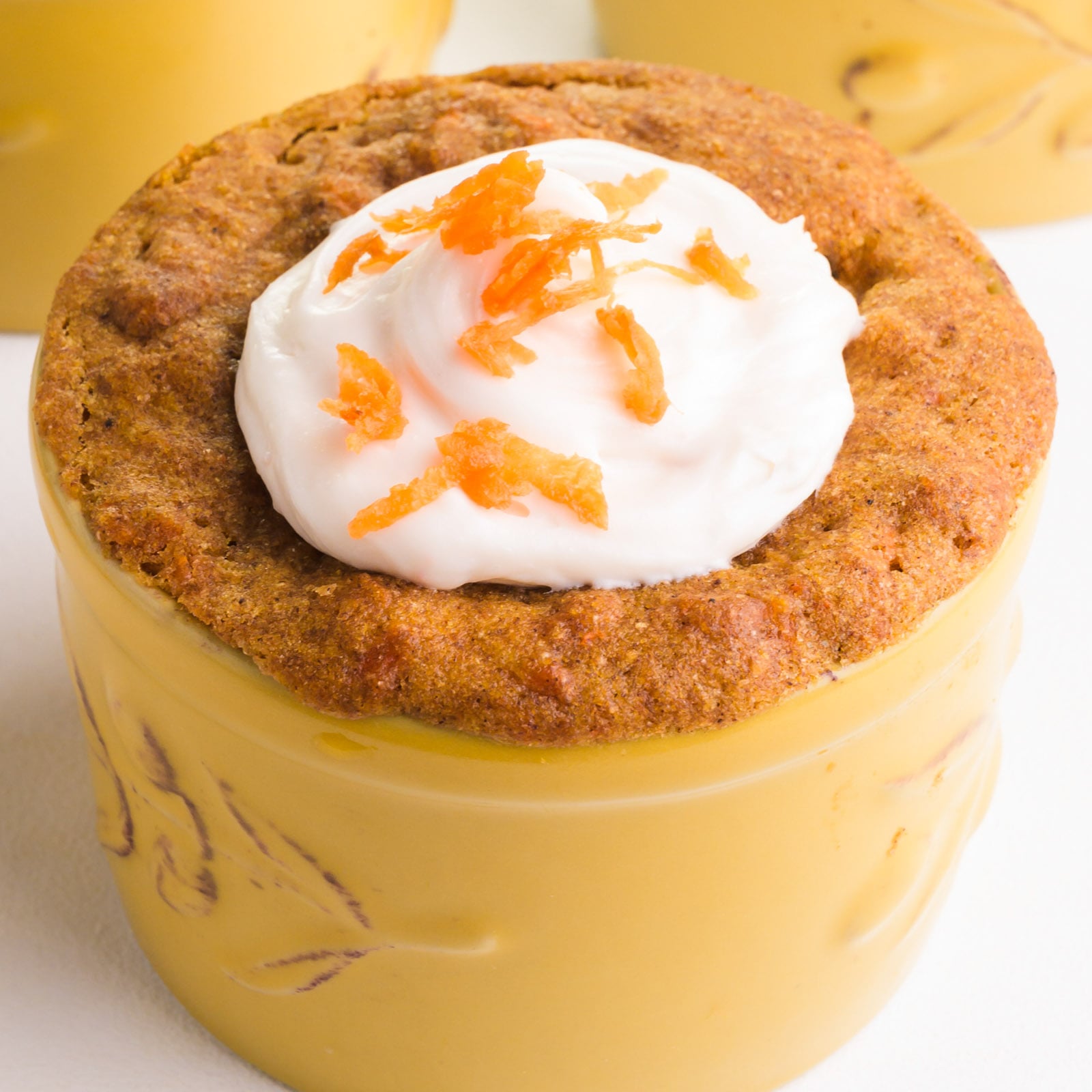 A closeup of carrot cake in a mug with cream cheese frosting and shaved carrot on top.