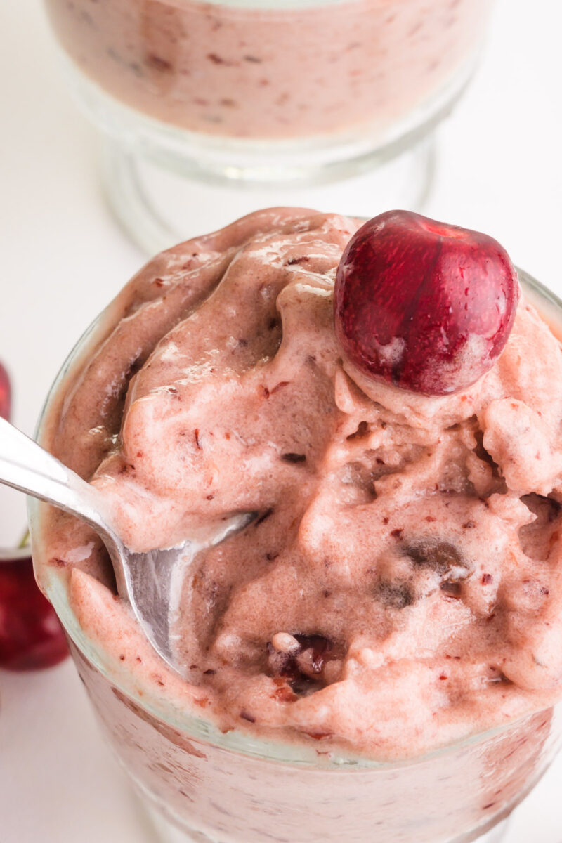 Looking down on a bowl of cherry banana ice cream with a spoon in it. There is a fresh cherry on top.