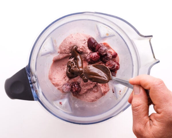 A hand drizzles spoonfuls of melted chocolate into a blender with a frozen cherry mixture.