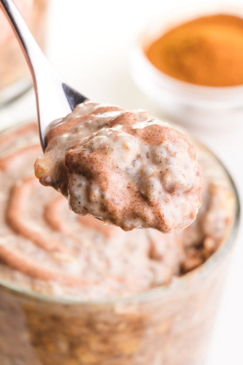A spoonful of overnight oats hovers over the rest of the bowl and a bowl of ground cinnamon in the background.