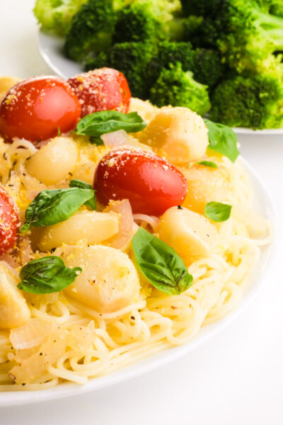 A plate of pasta has roasted garlic and tomatoes and fresh basil on top. There is steamed broccoli in the background.