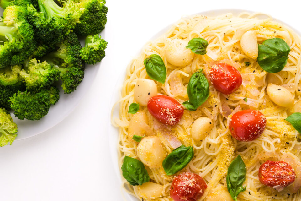 Looking down on a plate of pasta with tomatoes, garlic, and basil on top. There is a bowl of steamed broccoli beside it.