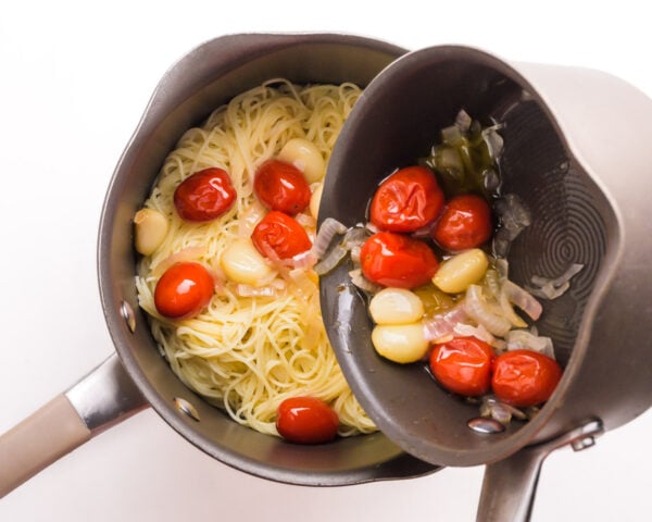 Roasted garlic, tomatoes, and onion, are being poured from one saucepan into another one with cooked pasta.