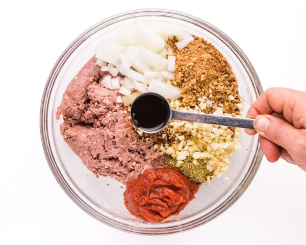 A hand holds a measuring spoon of sauce, pouring it over ingredients such as breadcrumbs and onions in a bowl.