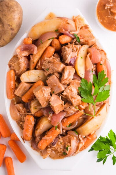 Looking down on a plant-based pot roast in a baking dish. It is surrounded by carrots, potatoes, herbs, and gravy.