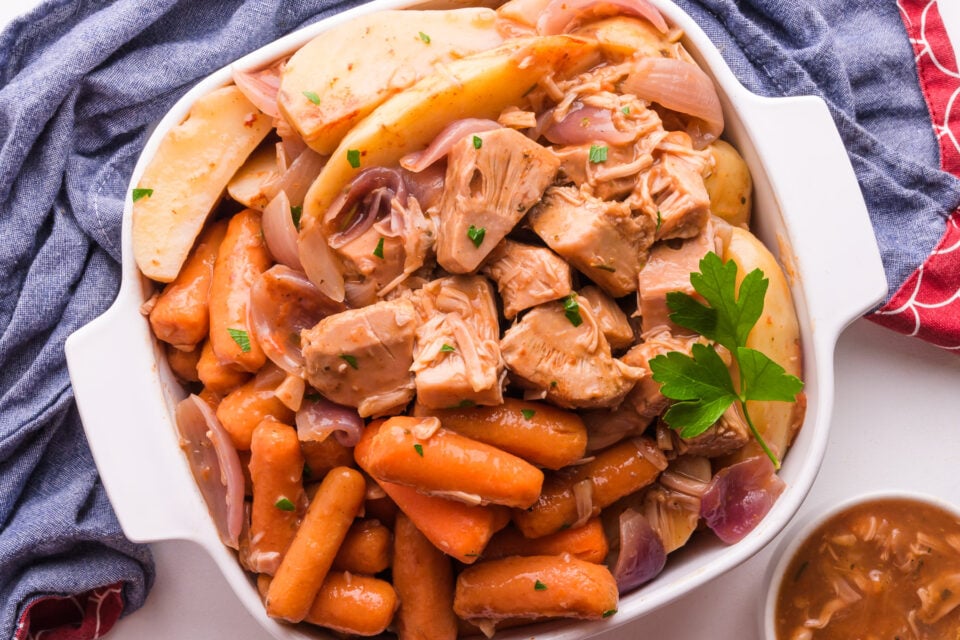 Looking down on vegan pot roast in a baking dish, with cooked carrots and potatoes. It is surrounded by a blue kitchen towel and some gravy in a bowl.