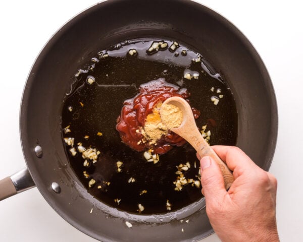 A hand holds a wooden measuring spoon, adding ingredients to a saucepan with garlic and sauce.