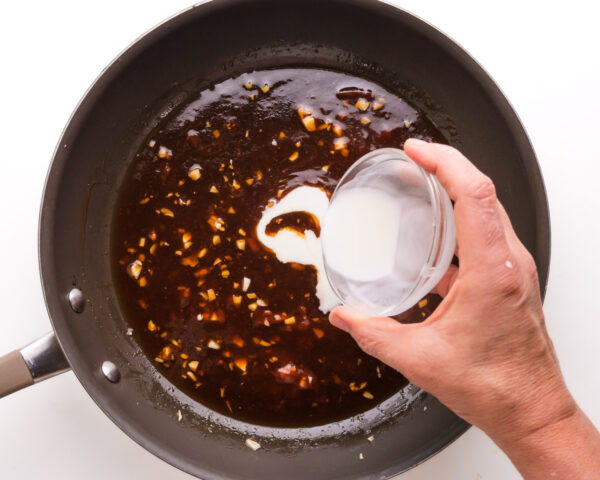 A hand holds a scowl bowl of cornstarch slurry, pouring it into a skillet with a sauce mixture.