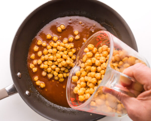 A hand holds a glass measuring cup full of chickpeas, pouring them into a skillet with sauce.