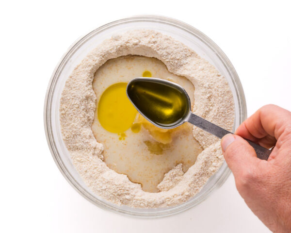 A hand pours a spoonful of olive oil into a bowl with other batter ingredients.