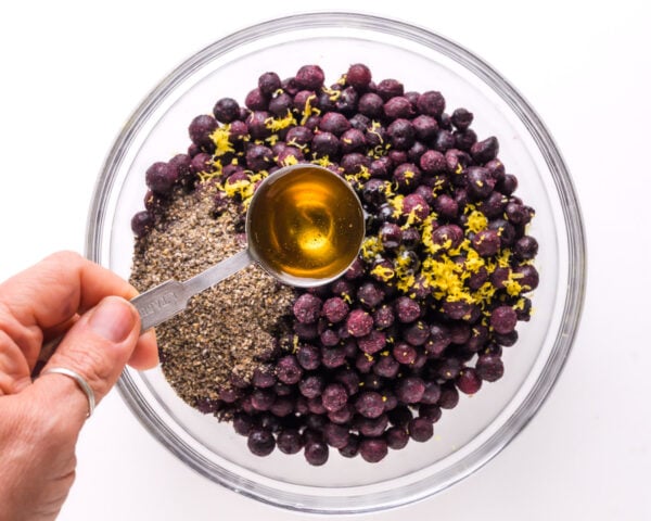 A hand holds a spoonful of maple syrup, pouring it into a bowl with blueberries, lemon zest, and more.