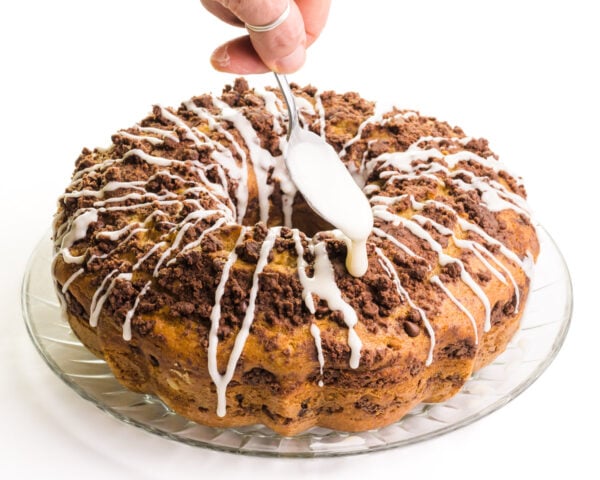 A hand holds a spoon drizzling vanilla icing over a coffee cake.