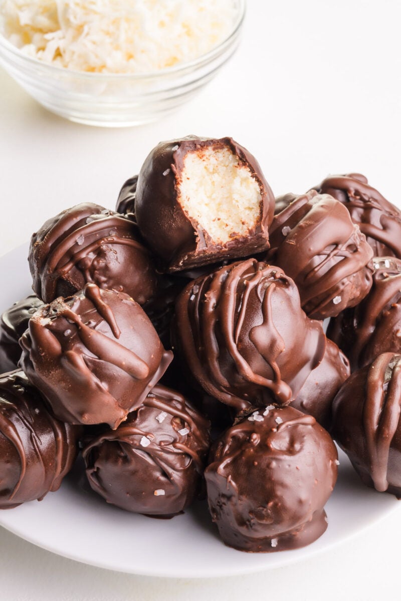 A stack of coconut balls sit on a plate. The top one shows a bite taken out. There is a bowl of coconut flakes in the background.