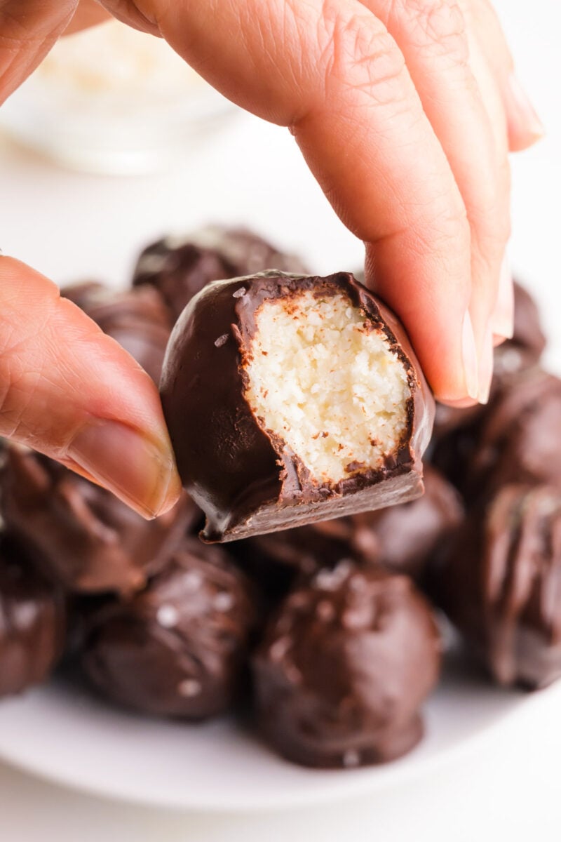 A hand holds a coconut ball with a bite taken out. It hovers over a plate with more of the treats.