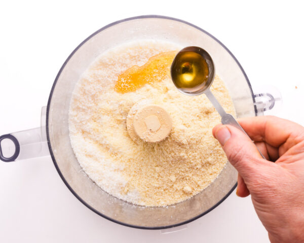 A hand holds a measuring spoon pouring syrup into a food processor with coconut flakes.