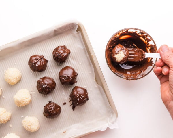 A hand holds a fork, dipping coconut balls in melted chocolate. There are more coconut balls on a pan, some are dipped others are not.