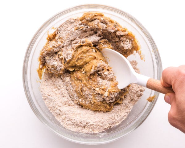 A hand holds a spatula stirring together flour ingredients with a wet mixture.