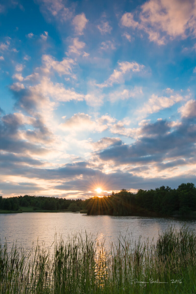 A sun sets over a lake, with colorful clouds in the sky and also reflected in the lake.