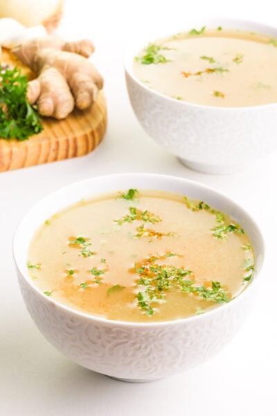 A bowl of biome broth sits in front of another bowl and a cutting board with ingredients like parsley and fresh ginger.