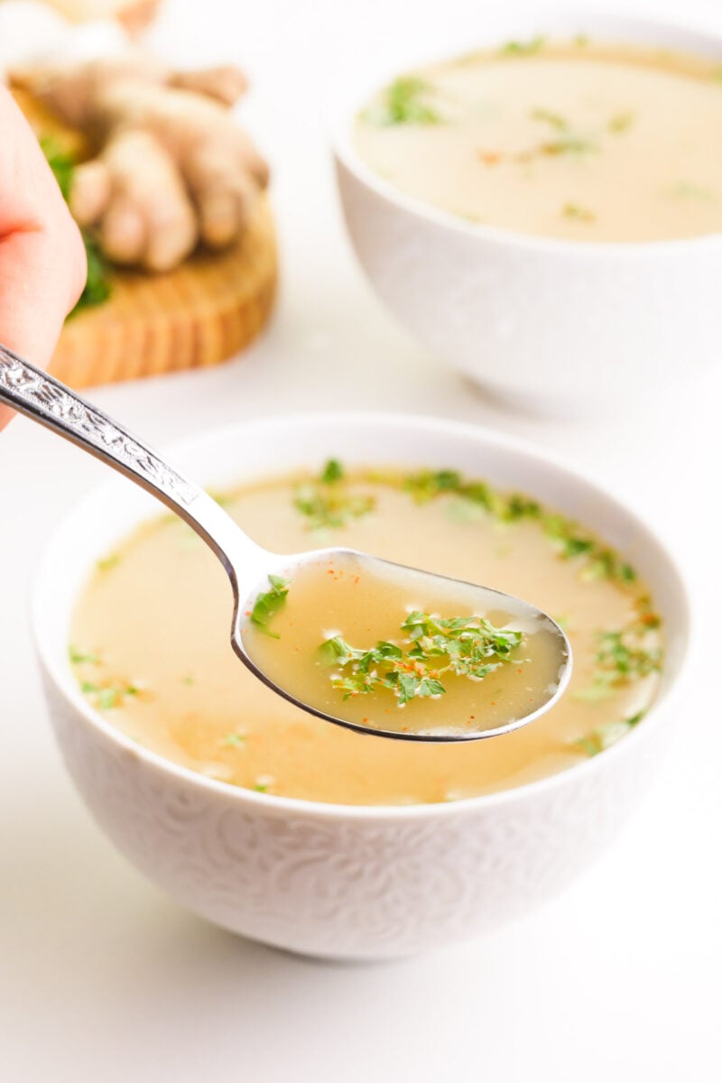 A spoonful of broth hovers in front of two bowls of soup and a cutting board with ingredients in the background.