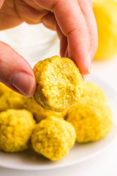 A hand holds a lemon energy bite with a bite taken out. There is plate with more energy balls and fresh lemons in the background.
