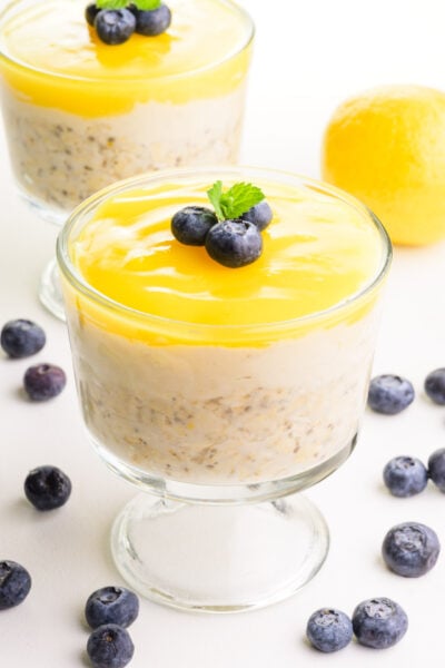 Two bowls of lemon overnight oats are surrounded by fresh blueberries and a fresh lemon in the background.