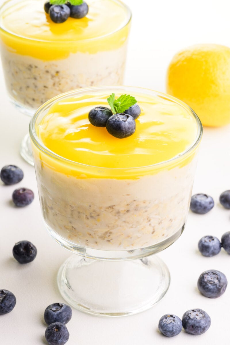 Two bowls of lemon overnight oats are surrounded by fresh blueberries and a fresh lemon in the background.