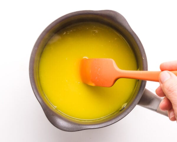 A hand holding an orange spatula stirring bright yellow liquid in a dark saucepan on a white background.