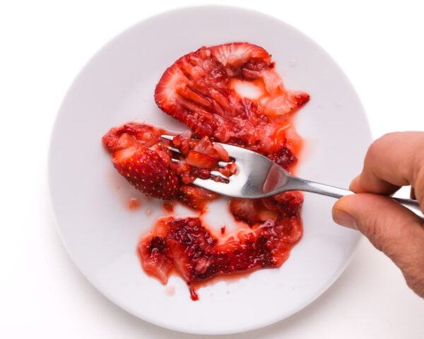 A hand holds a fork, mashing strawberries on a plate.