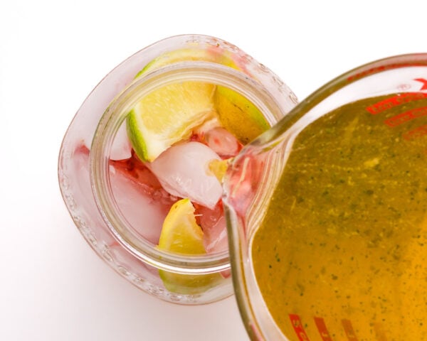 Green tea is being poured from a pyrex measuring cup into a mason jar with ice, mashed strawberries, and, lime slices.