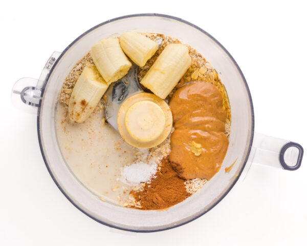 Looking down on a food processor bowl with ingredients such as sliced bananas, peanut butter, spices, and more.
