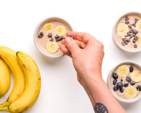 A hand drops toppings onto batter in a ramekin. There are two more batter-filled ramekins beside it along with whole bananas.