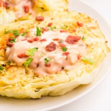 Cabbage steaks are sitting on a plate. They are topped with dressing and veggie bacon bits.