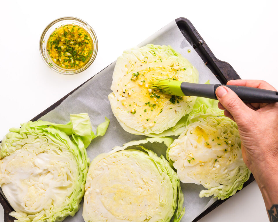 A hand brushes dressing over cabbage steaks on a prepared pan.