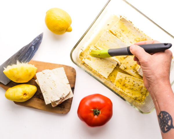 A hand holds a pastry brush, brushing marinade on tofu slices in a small dish. There are lemons and tomatoes and a cutting board nearby.
