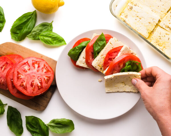 A hand places tofu on a plate with sliced tomatoes and basil leaves. There is more tofu in a dish, fresh basil leaves, a lemon, and sliced tomatoes nearby.