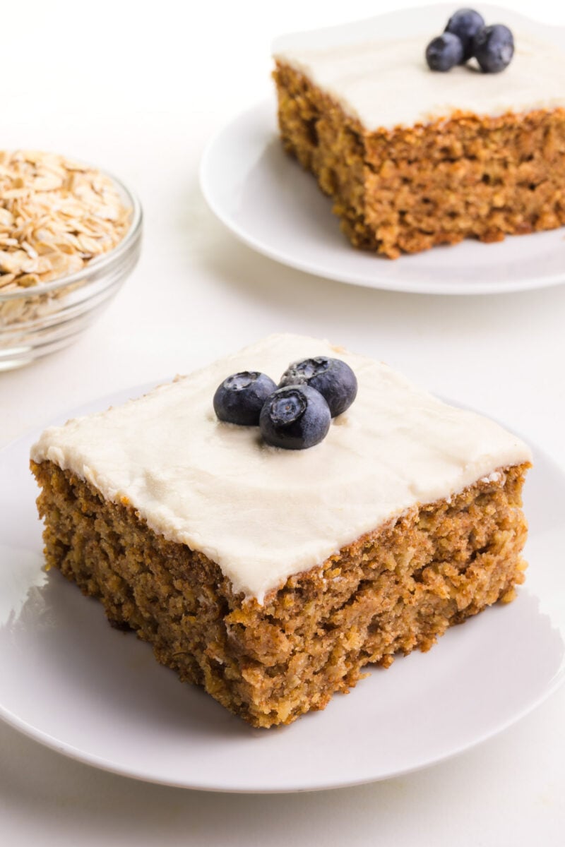 A slice of cake on a plate sits infant of a bowl of oats and another slice in the background.