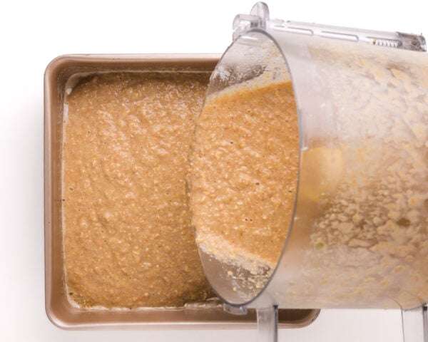 Cake batter is being poured from a food processor into a square cake pan.