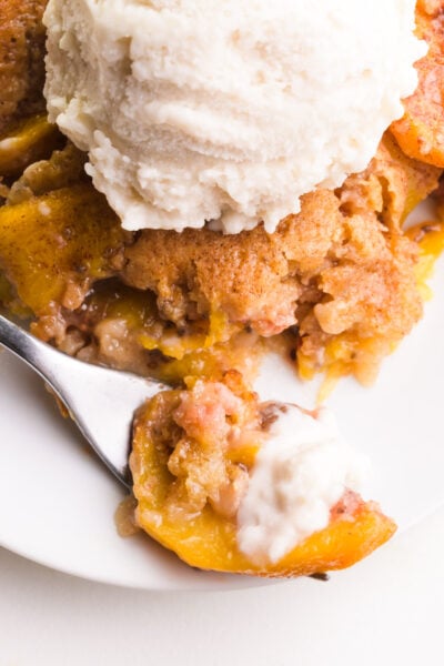 A forkful of vegan peach cobbler sits in front of the rest of the cobbler on a plate. It's topped with vegan vanilla ice cream.